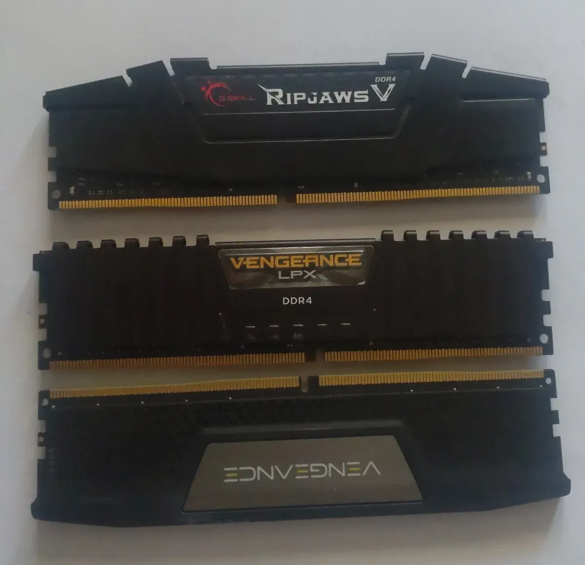 Three RAM sticks. Two topmost are DDR4 and the one at the bottom is DDR5. The two at the bottom are positioned so the pins are lying opposite to each other. On each stick there is a visible cutout at the bottom where pins are located. There is a noticeable difference in positioning the cutout between DDR4 and DDR5.